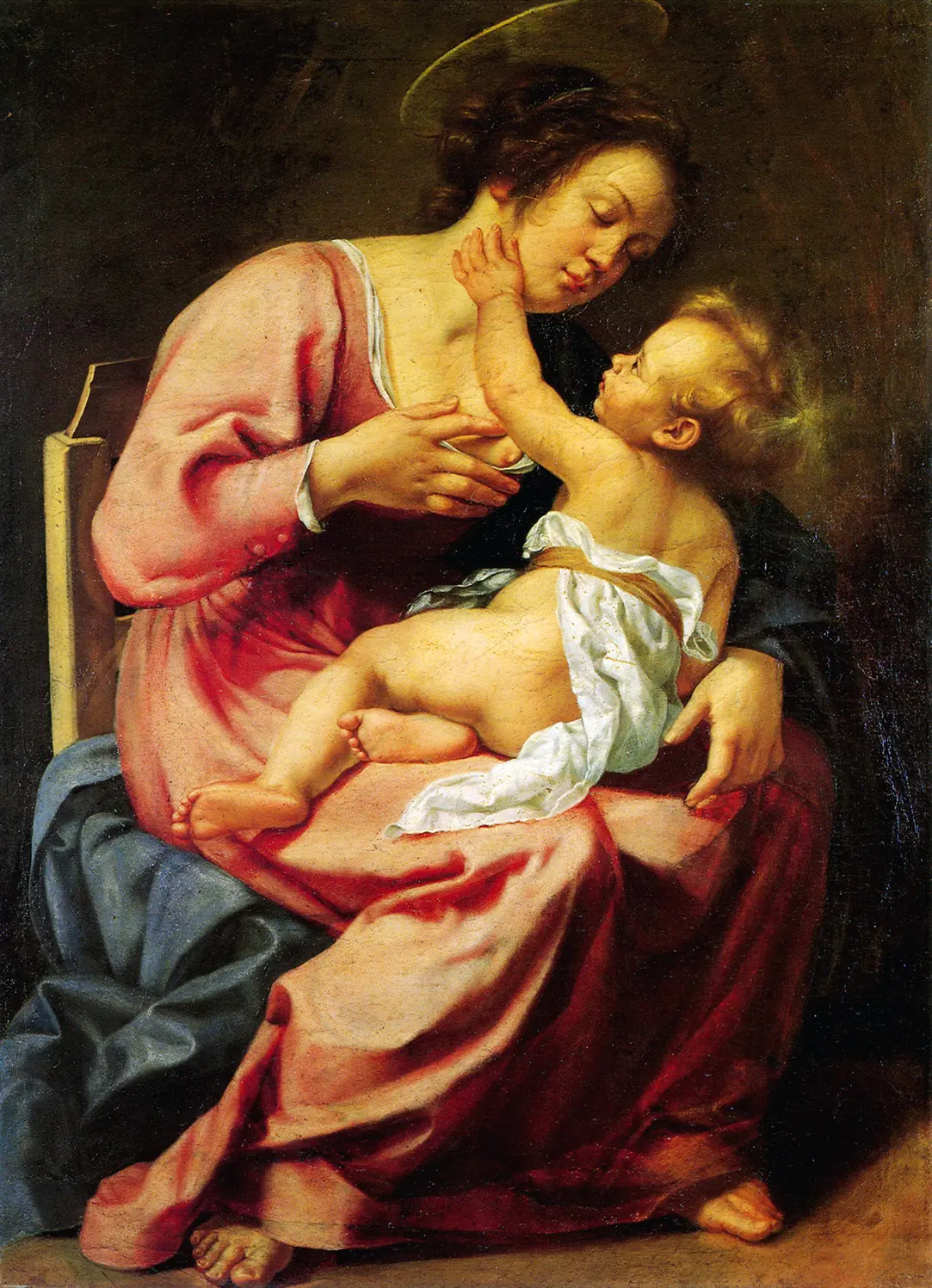 Seated mother in a pink dress holds her baby on her lap as he reaches up to touch her chin