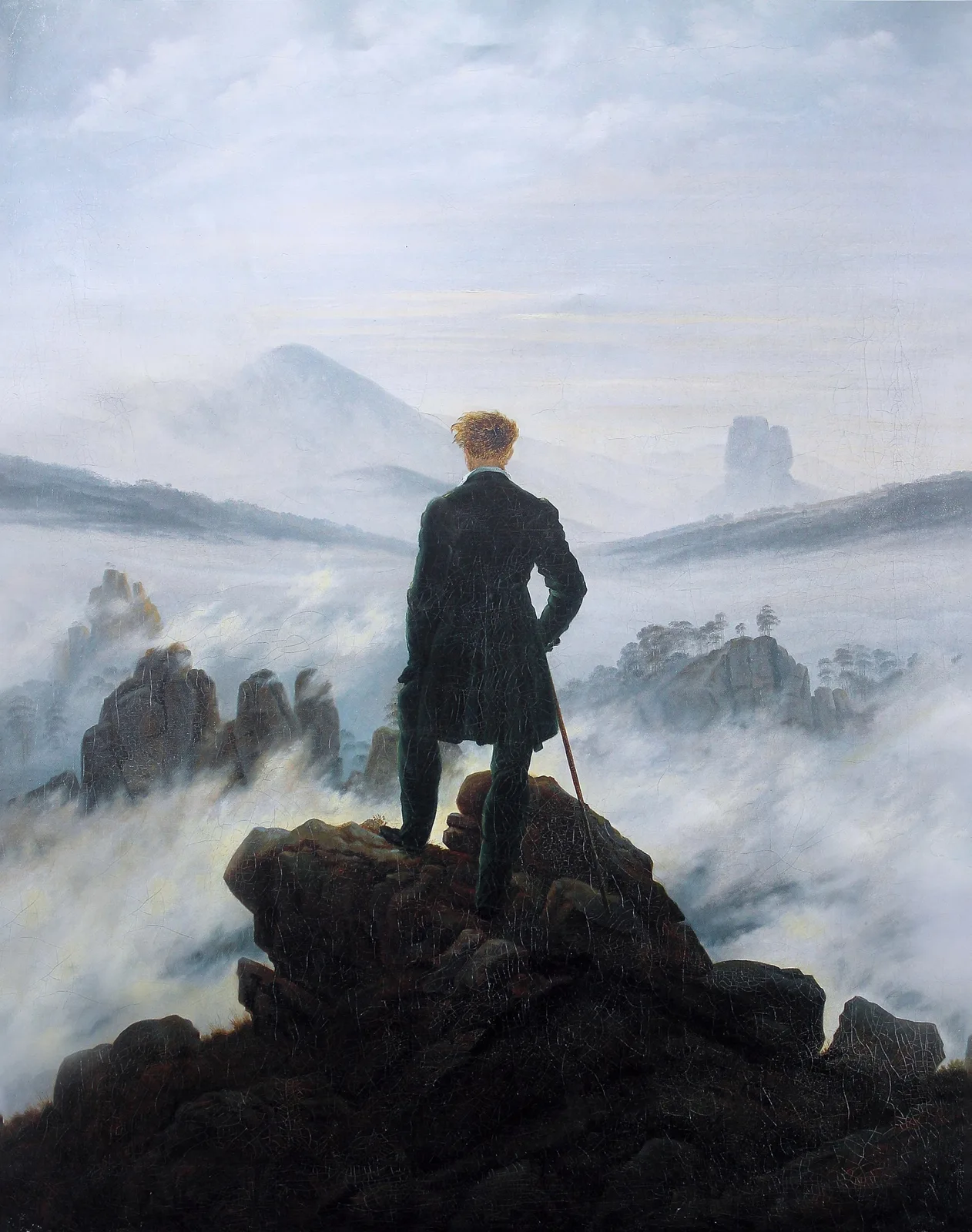 Hombre de espaldas, con abrigo oscuro y bastón, de pie sobre una roca frente a un mar de niebla y montañas lejanas