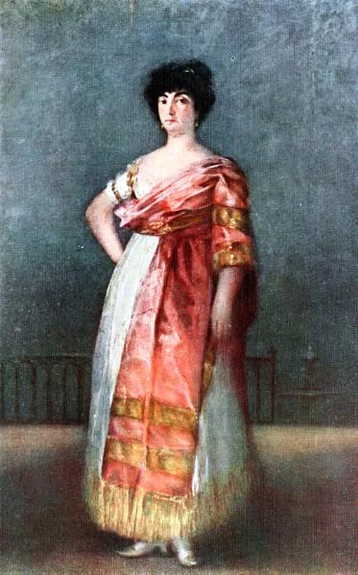 Mujer de pie con una mano en la cadera, cabello oscuro corto y chal rosado con franjas doradas sobre falda blanca