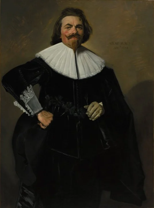 Hombre de pie vestido de negro, con gran cuello blanco, una mano en la cadera y un guante en la otra, sonriendo