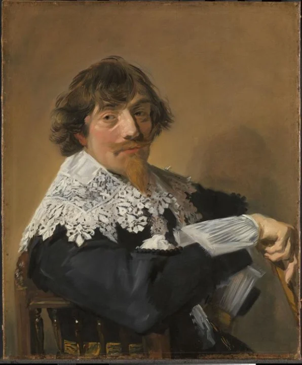 Hombre de cabello castaño y barba rojiza, sentado de lado, apoya el brazo en el respaldo de la silla y voltea a mirarnos