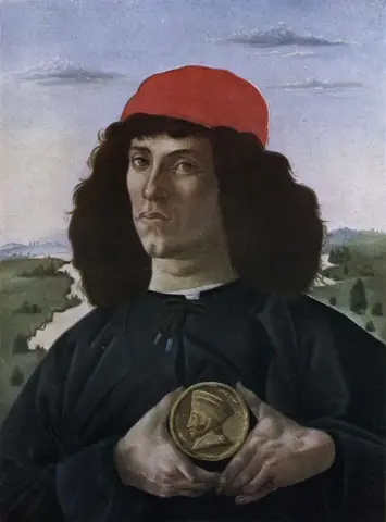 A young man in a red cap with long dark hair holds a gold medal in both hands, against a landscape with a river