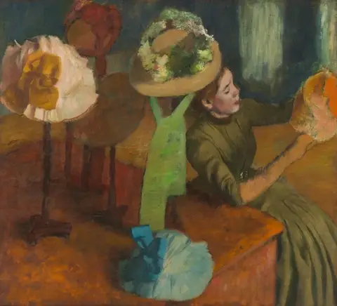 Mujer de vestido verde olivo que sostiene un sombrero anaranjado, junto a sombreros de colores en sus bases