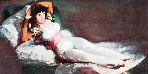 Woman reclining on a green couch against white pillows, in a close-fitting white dress, pink sash and orange jacket