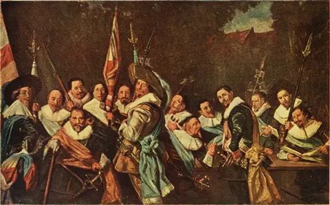 Grupo de oficiales con cuellos blancos, bandas de colores, picas y banderas, reunidos al aire libre junto a una mesa