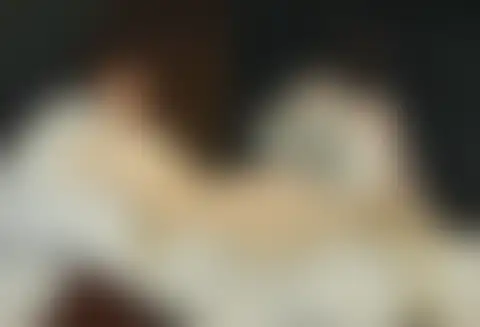 Mujer desnuda recostada en una cama blanca; detrás, otra mujer con vestido rosa le acerca un ramo de flores