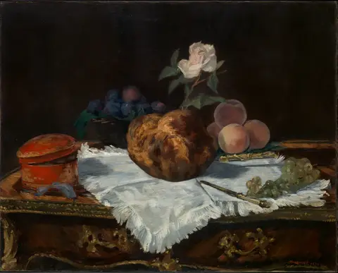 Bollo dorado con una rosa blanca encima, sobre una servilleta blanca, con duraznos, uvas, ciruelas y una caja roja