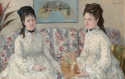 Dos jóvenes con vestidos blancos de lunares azules sentadas en un sofá floreado; una sostiene un abanico abierto