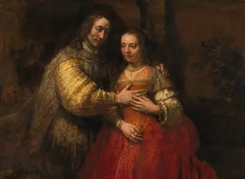 Pareja muy junta: él, de mangas doradas, apoya la mano en el pecho de ella, vestida de rojo, y ella la cubre con la suya