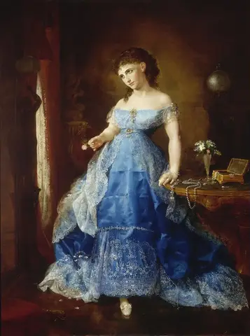 Standing woman in a long blue satin gown with white lace, holding a rose beside a table strewn with pearls