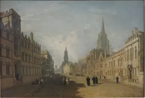 Calle ancha de Oxford con edificios de piedra a ambos lados, una aguja gótica alta y estudiantes con togas negras