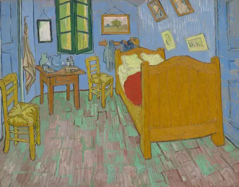 Cuarto de paredes azules con cama de madera amarilla, cobija roja, dos sillas de paja y cuadros colgados en la pared