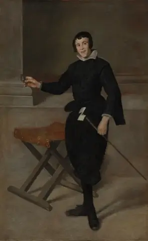 Smiling young man in black holding something small in one hand and a long stick in the other, beside a folding stool