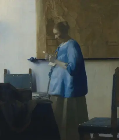 Mujer de chaqueta azul, de pie y de perfil, lee una carta ante un mapa colgado, entre una mesa y dos sillas