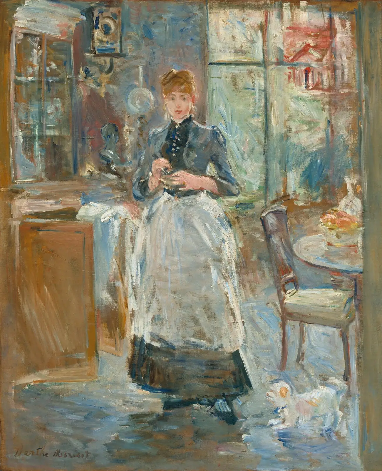 Young woman in a dark blue blouse and white apron standing in a dining room by a cabinet, a chair and a window