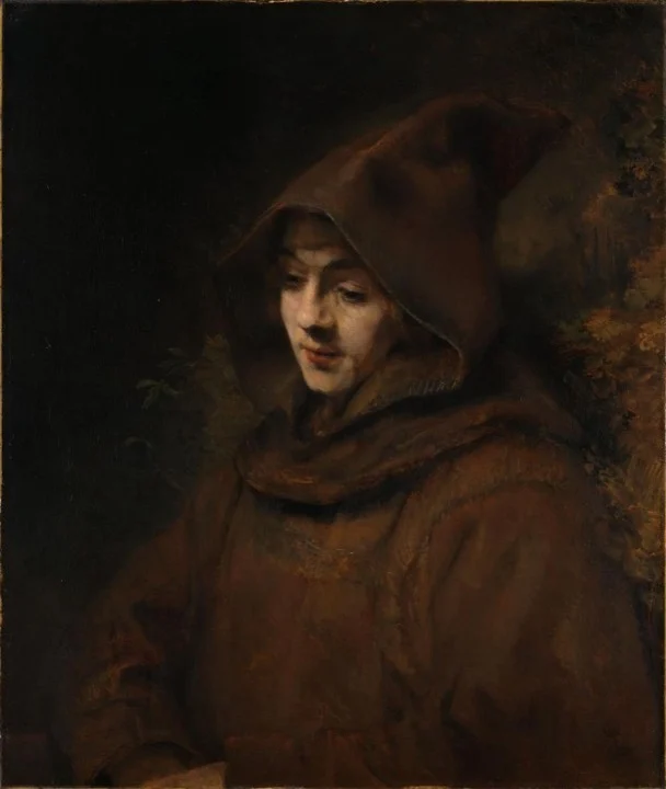 Young man in a brown habit with a pointed hood, eyes lowered and a faint smile, against dark foliage